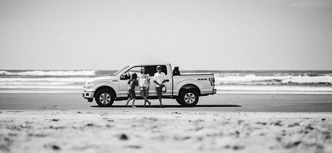 A cinematic black and white photograph of Vince Chapple and his sons standing beside a pickup truck on Long beach during a road trip through the American West.